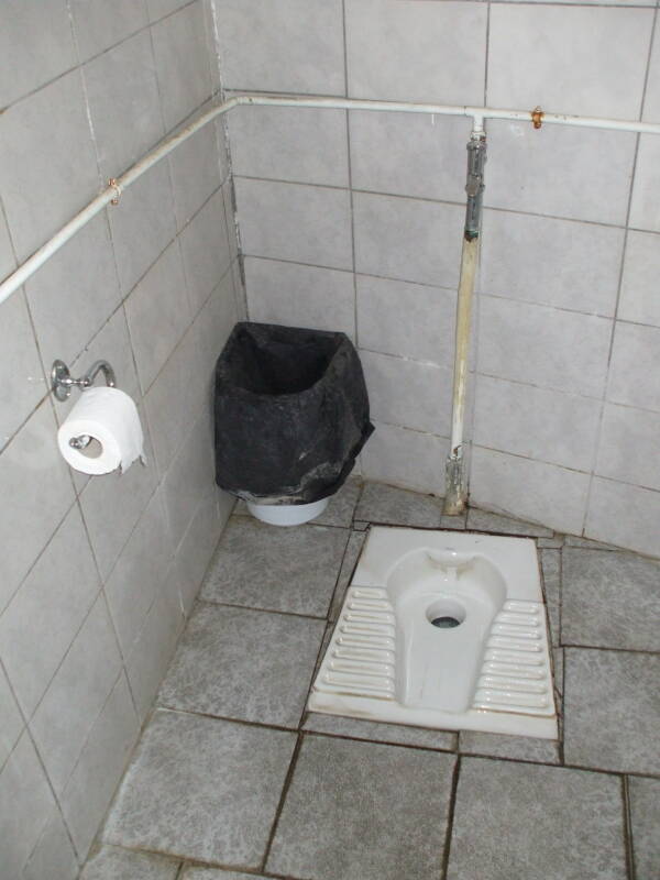 Greek squat toilet in a monastery at Meteora, with toilet paper roll, waste bin, and visible plumbing.