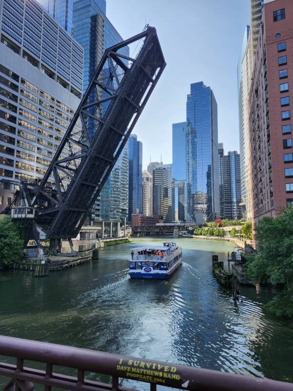 North Branch of the Chicago River seen from Kinzie Street Bridge; an architectural tour boat continues to the south under a raised bridge. In the distance, the South Branch continues ahead, the main Chicago River stream runs east, to the left, into Lake Michigan. An El train crosses the river in front of skyscrapers.