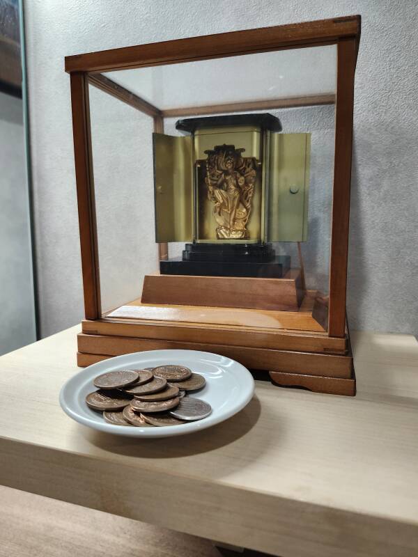 Small statue of Ucchuṣma in the bathroom at the temple, in a glass case with a dish of coins in front of it.