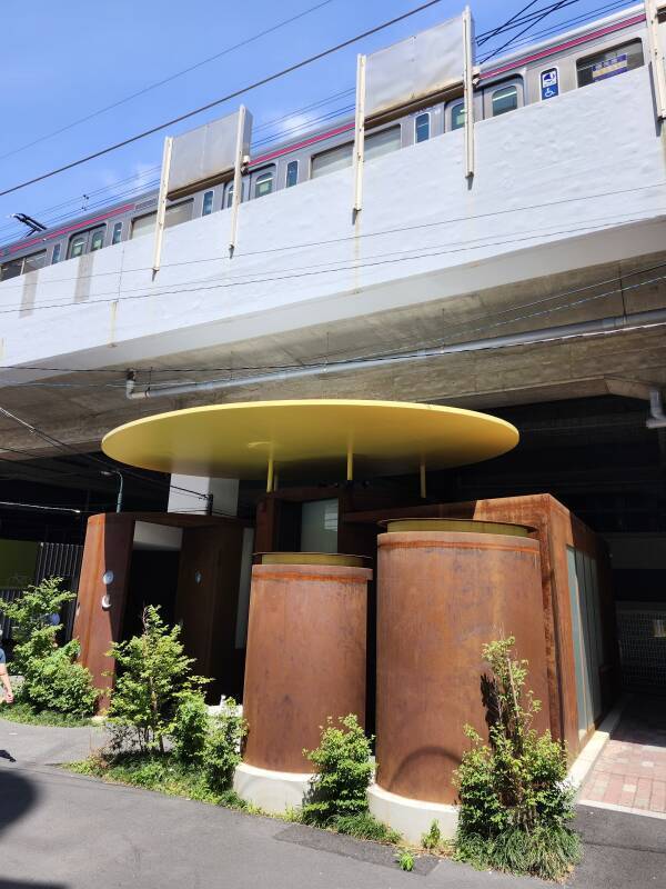 Sasazuka Greenway Public Toilet: oxidized steel cylinders arranged beneath a yellow awning, all of it partially under an elevated rail line.